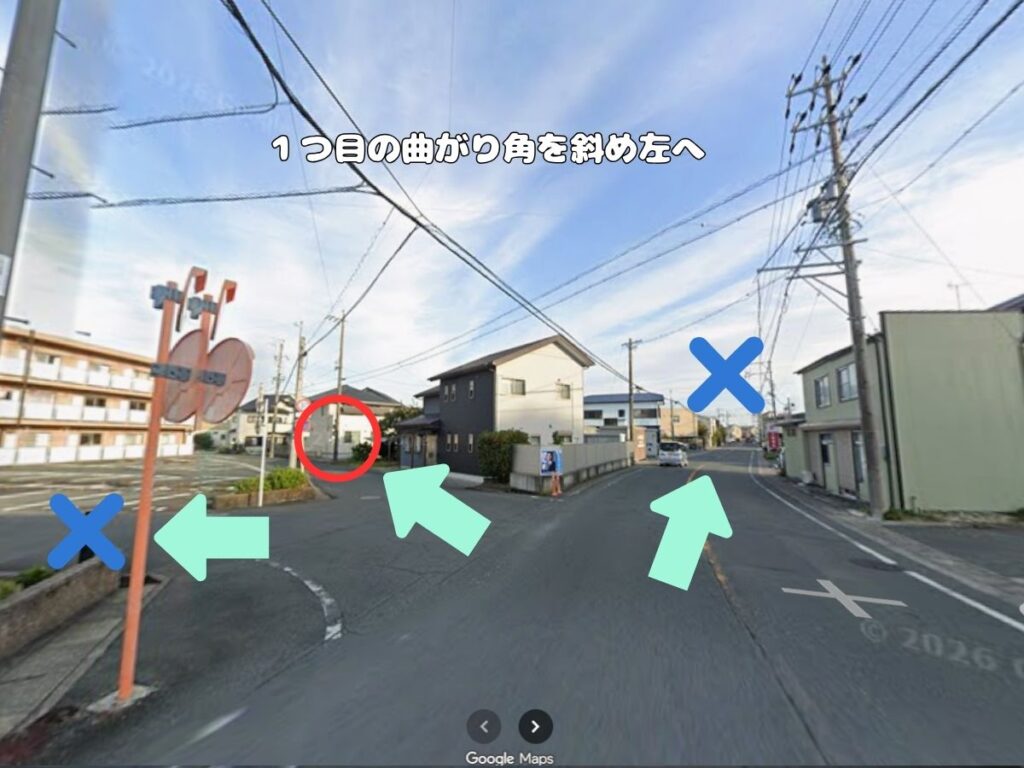 トリミングサロン「ドッグアシル」からの帰り道案内。「1つ目の曲がり角を斜め左へ」のテロップが入り、誤進入を防ぐ青い×印と、進路を示すミントグリーンの矢印、赤丸で囲まれた目印がある三差路の風景写真